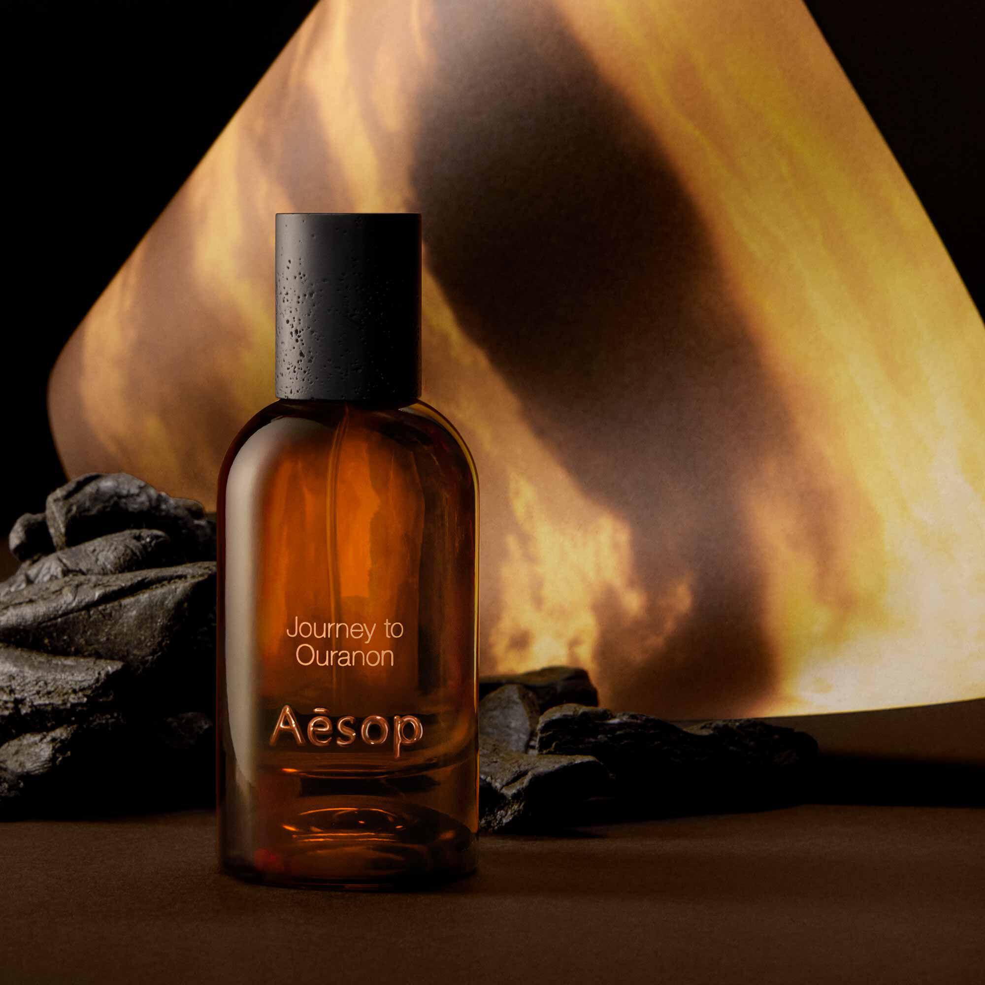 Aesop Journey to Ouranon bottle beside dark stones and a glowing orange backdrop.