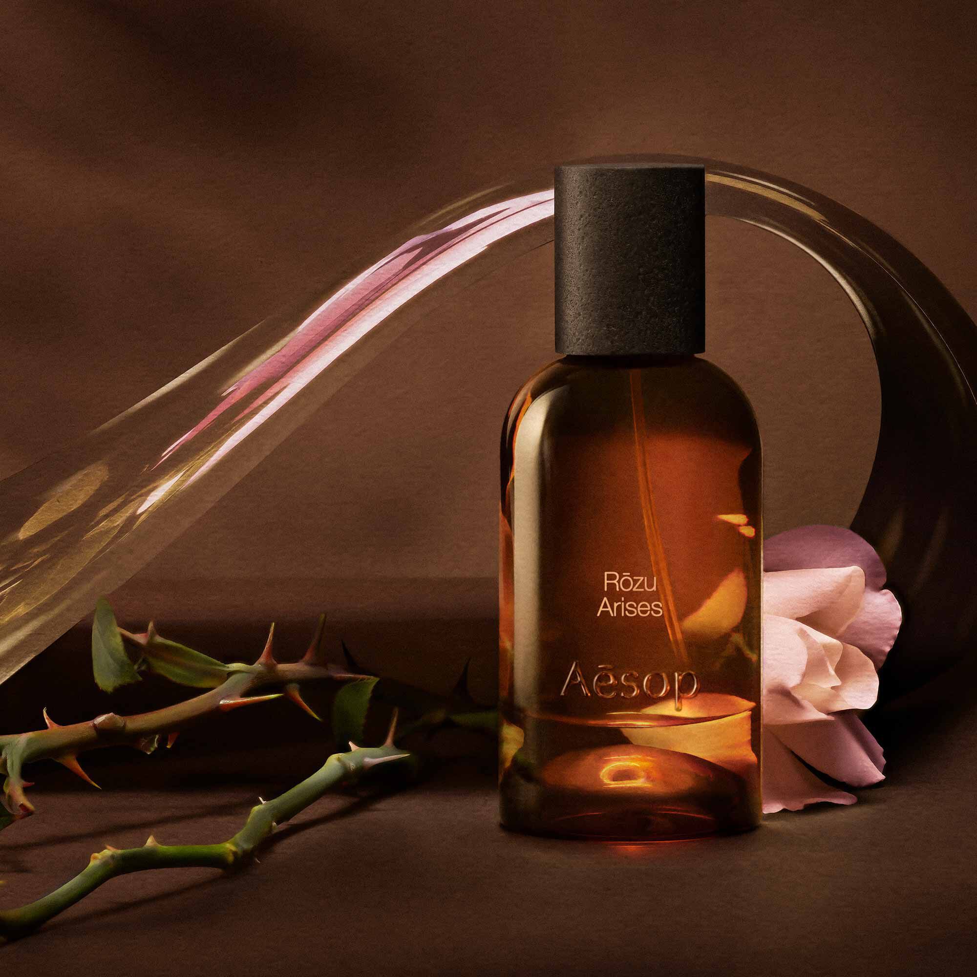 Aesop Rozu Arises bottle with a rose stem and flower petals on a warm surface.
