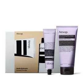 Two lilac Aesop tubes with black caps stand in front of a silver gift box with a gold illustrated sleeve.
