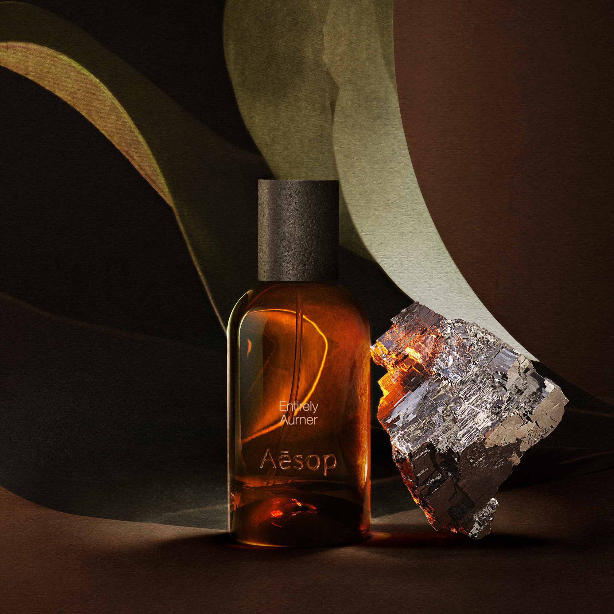 Aesop Entirely Aurner bottle beside a mineral fragment and broad green leaves.