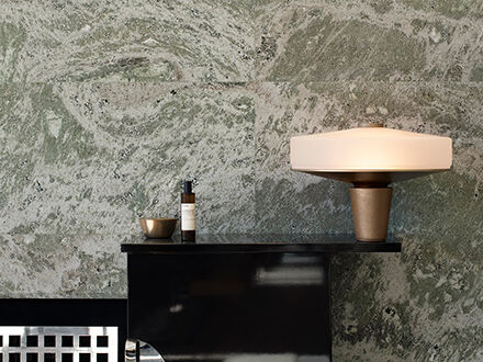 A wide, glowing modern table lamp with a bronze base sits on a black sideboard against a green marbled stone wall. A small gold bowl and a dark bottle are also on the sideboard.