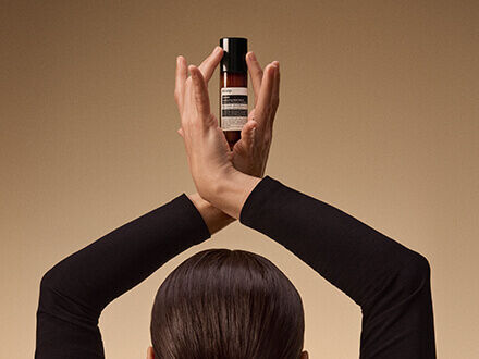 A person in black clothing with their back to the camera, holding a small amber bottle above their head with both hands.