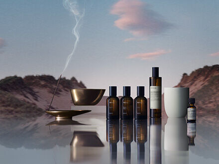 A collection of Aesop home fragrance products, including amber glass spray bottles, an oil burner, and incense, arranged on a reflective surface against a serene desert landscape at dusk.