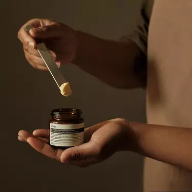 An editorial, mid-shot photograph of a person holding an open amber glass jar of Aesop "Perfect Facial Hydrating Cream" in one palm. With their other hand, they use a sleek silver metal spatula to scoop out a small dollop of the thick, pale-yellow cream. The person is wearing a minimalist, earth-toned brown apron against a soft, dark-olive textured background.