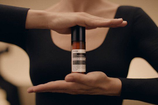 A person's hands holding a bottle of Aesop Solais Replenishing Hand Serum vertically against a warm, minimalist background.