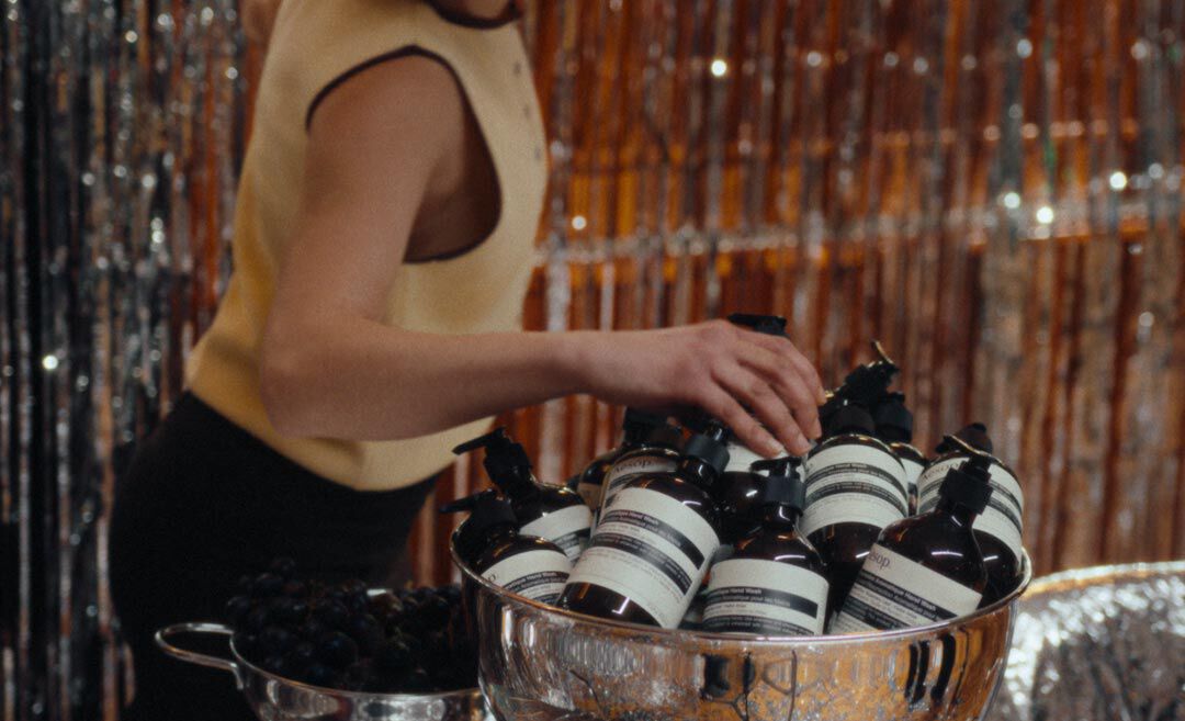 Person in a pale yellow sleeveless top reaching towards Aesop pump bottles piled in a large silver bowl, with tinsel behind.