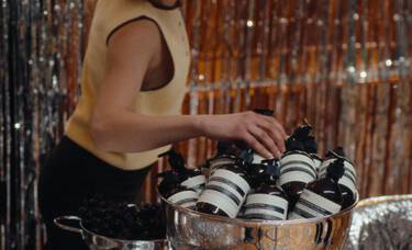 Person in a pale yellow sleeveless top reaching towards Aesop pump bottles piled in a large silver bowl, with tinsel behind.