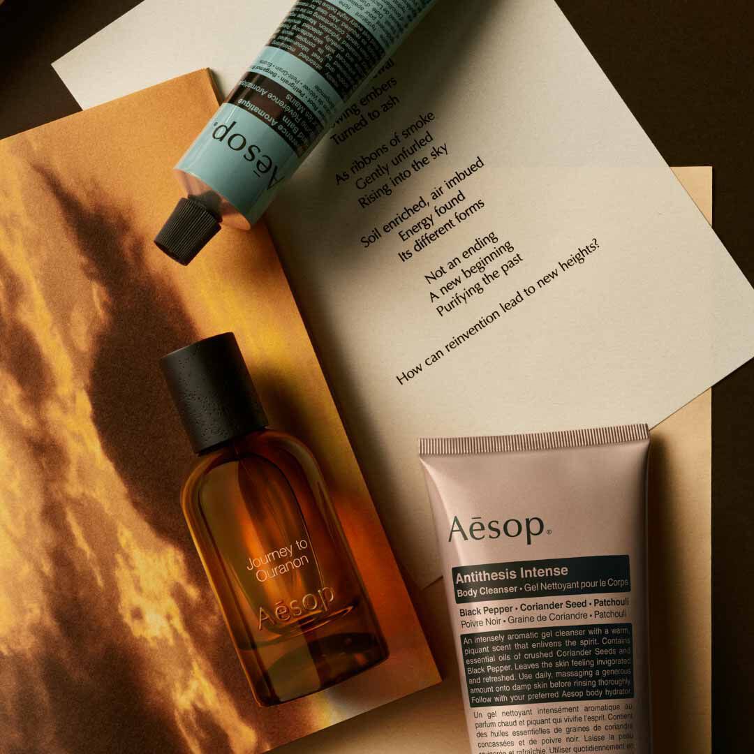 Aesop Journey to Ouranon fragrance bottle, cosmetic tube and printed page on a surface with a golden sky backdrop.