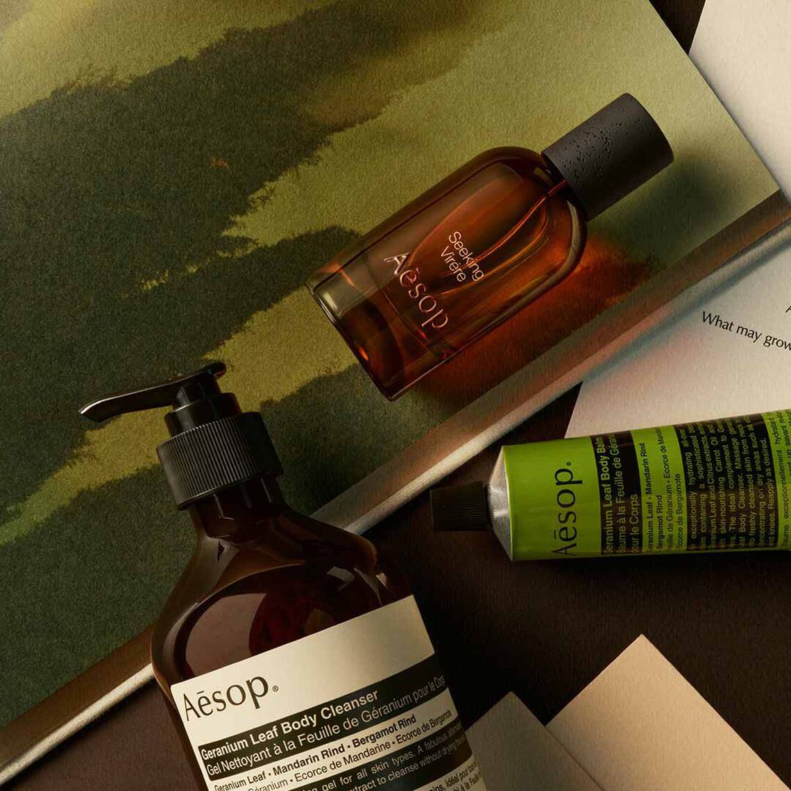 Aesop Seeking, Virere fragrance bottle, large pump bottle and cosmetic tube on an open book on a green textured background.