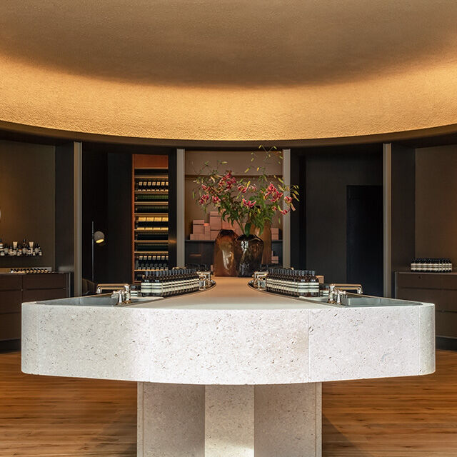 An elegant beauty boutique with minimalist design, featuring a large stone central island with integrated sinks and products, surrounded by dark wood shelving and warm ambient lighting.