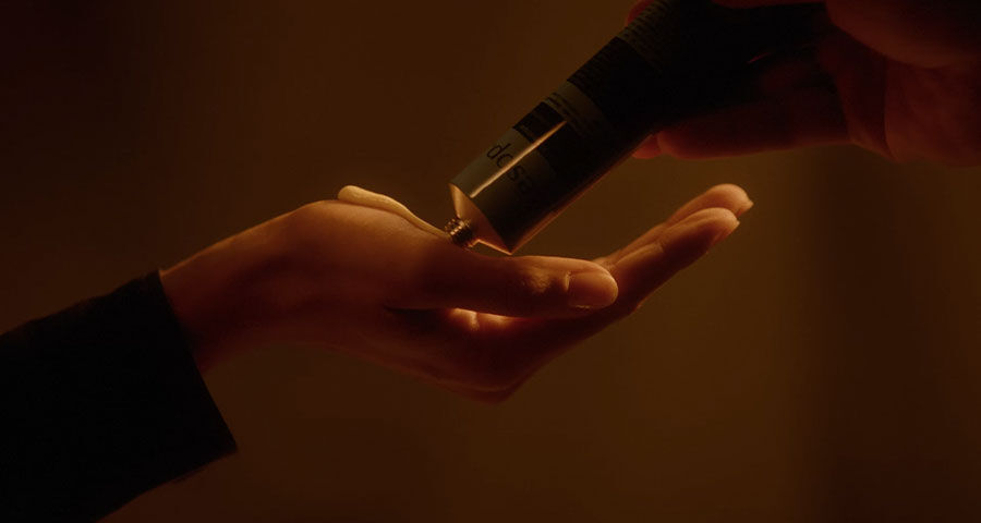 A hand squeezes a black tube, dispensing a dollop of cream onto another hand, against a dark, warm-toned background.