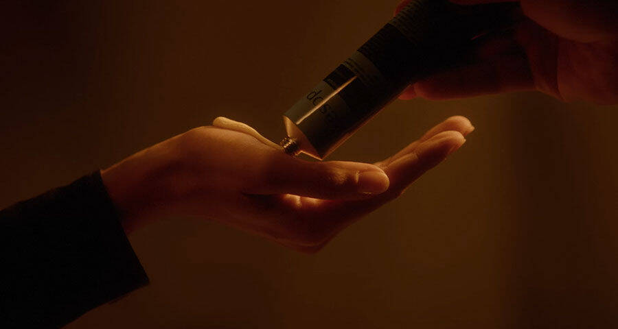A hand squeezes a black tube, dispensing a dollop of cream onto another hand, against a dark, warm-toned background.