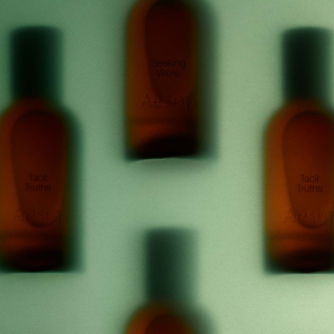 An atmospheric, blurry close-up view of amber-tinted Aesop bottles behind a frosted or backlit surface, with faint text reading "Tacit Truths" and "Aesop".