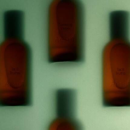 An atmospheric, blurry close-up view of amber-tinted Aesop bottles behind a frosted or backlit surface, with faint text reading "Tacit Truths" and "Aesop".