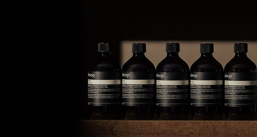 A row of six identical dark Aesop bottles with black labels and caps are neatly lined up on a textured brown surface against a dark background.