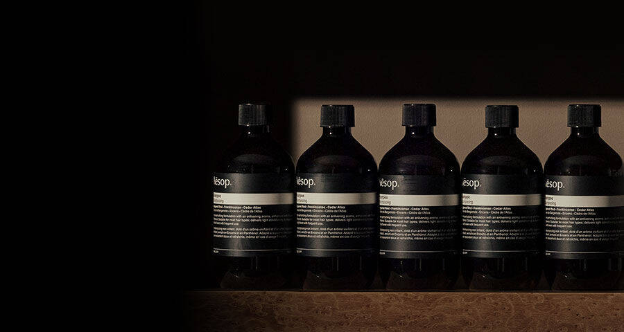 A row of six identical dark Aesop bottles with black labels and caps are neatly lined up on a textured brown surface against a dark background.