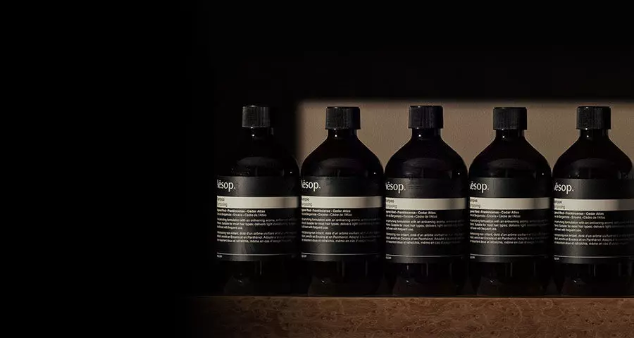 A row of six identical dark Aesop bottles with black labels and caps are neatly lined up on a textured brown surface against a dark background.