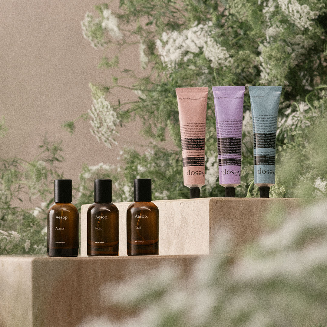 A still-life product photo featuring three Aesop amber glass perfume bottles and three colorful hand cream tubes arranged on stone pedestals amidst lush green foliage.