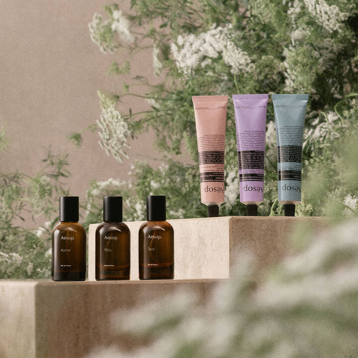 A still-life product photo featuring three Aesop amber glass perfume bottles and three colorful hand cream tubes arranged on stone pedestals amidst lush green foliage.
