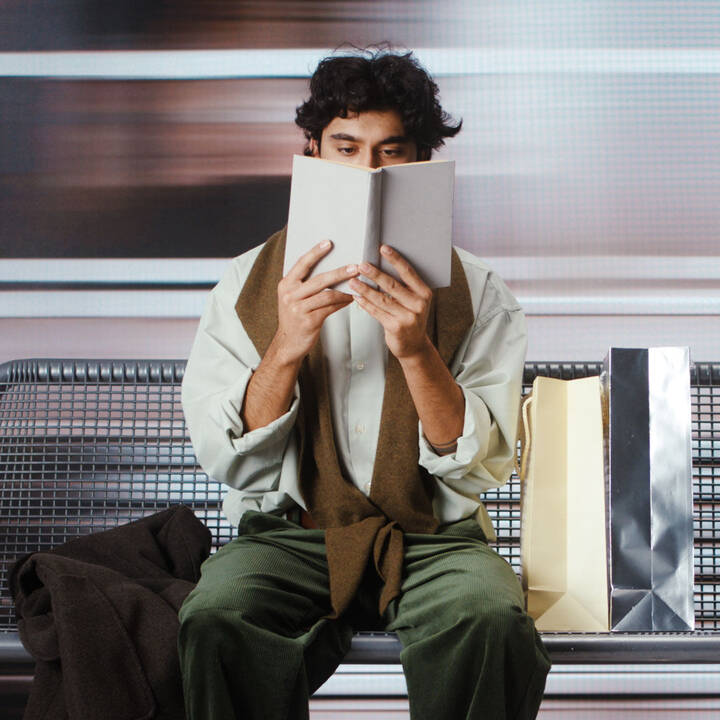 Person reading a book on a metal bench beside shopping bags, wearing a pale green shirt and green trousers, with blurred movement behind.