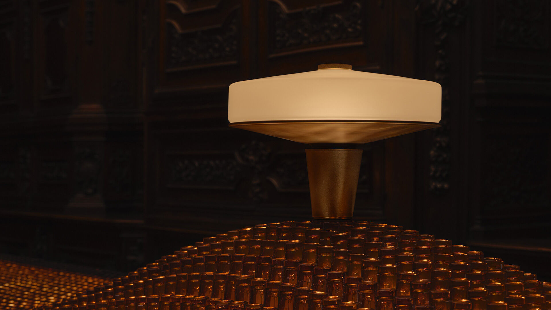 A minimalist modern lamp sitting atop a mound of amber glass bottles against a dark, ornate wood-carved background.