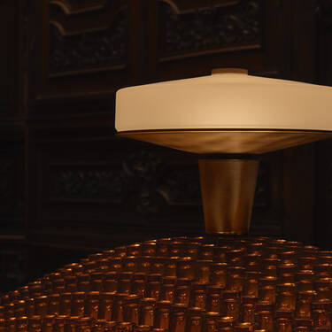 A minimalist modern lamp sitting atop a mound of amber glass bottles against a dark, ornate wood-carved background.