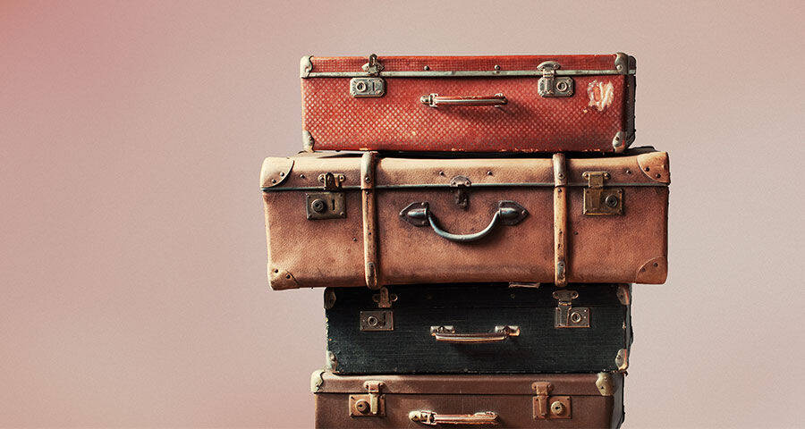 A stack of four vintage suitcases in various shades of brown and red is set against a soft, gradient background of pink and peach.