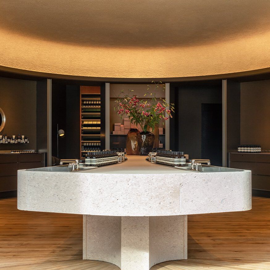 An elegant beauty boutique with minimalist design, featuring a large stone central island with integrated sinks and products, surrounded by dark wood shelving and warm ambient lighting.
