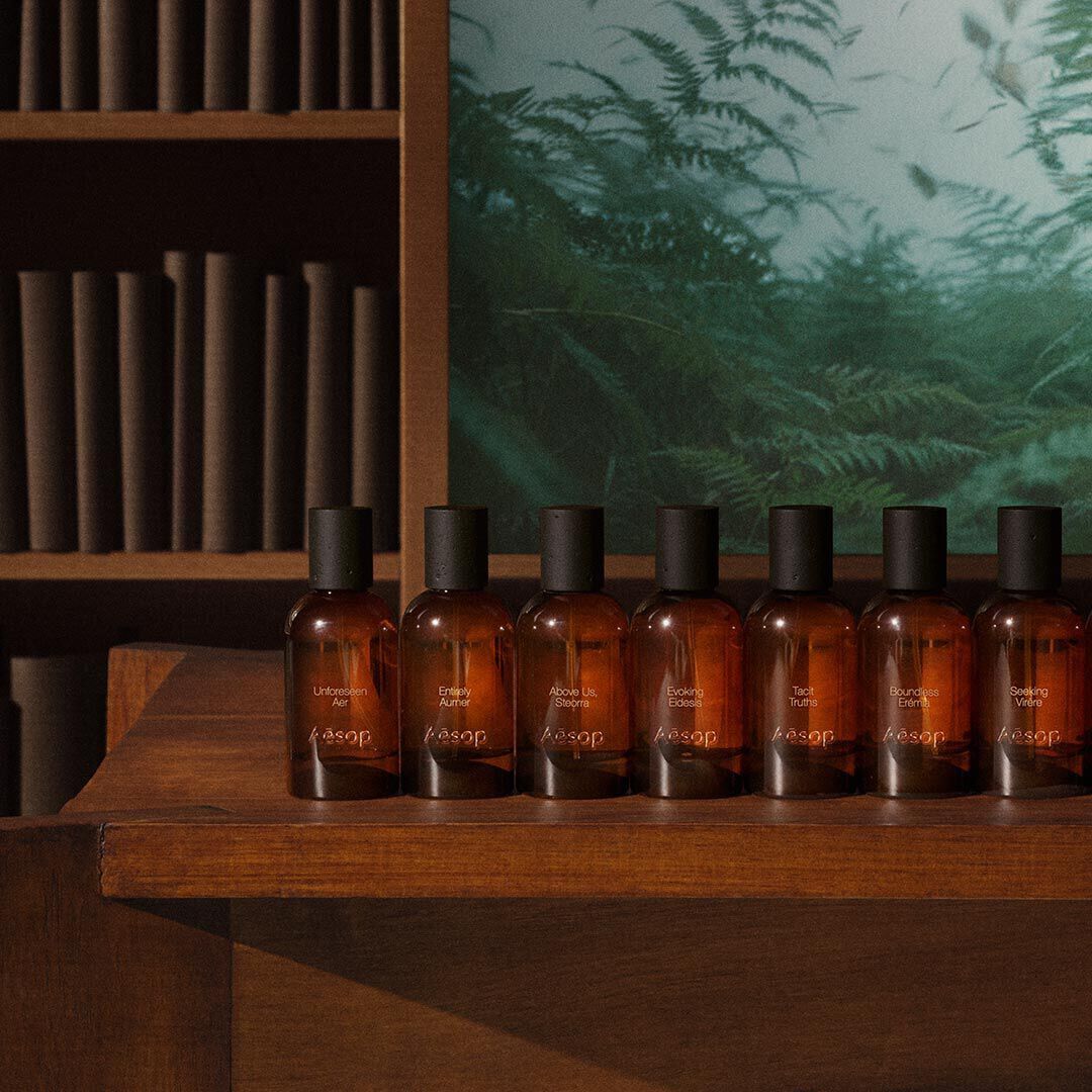 Row of Aesop amber fragrance bottles with black caps lined up on a dark wooden shelf, with cylindrical wooden elements on shelves and a green fern panel in the background.
