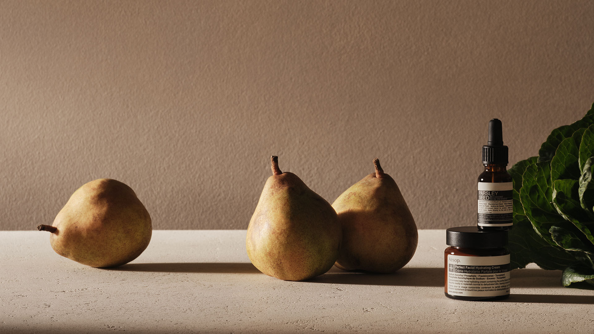A minimalist still life featuring three pears, green leafy vegetables, and stacked Aesop skincare bottles on a textured beige surface against a warm, neutral background.