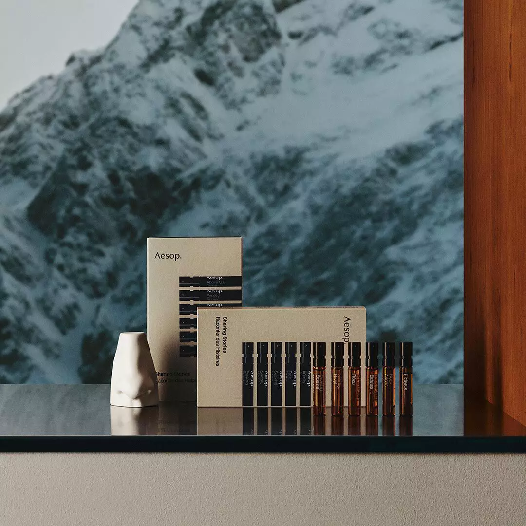 Two Aesop boxes, six small amber vials and a white nose-shaped object sit on a reflective ledge overlooking snow-covered mountains.