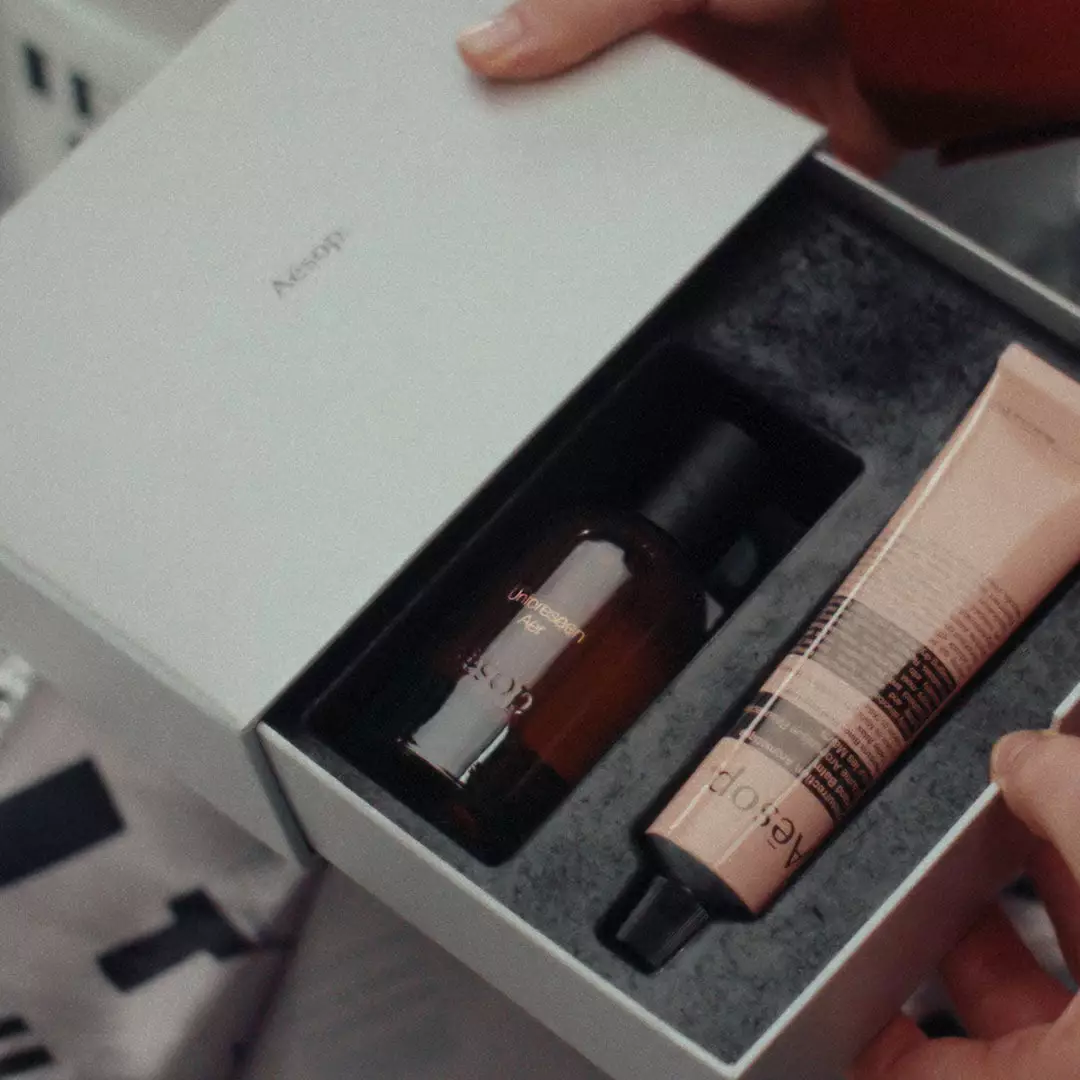 Hands hold an open silver Aesop box containing an amber bottle and a pink tube nestled in a dark fitted insert.