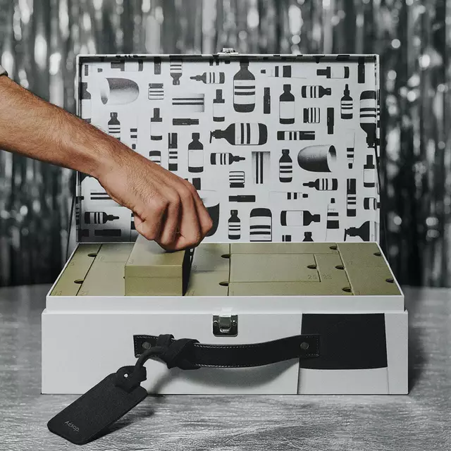 A hand lifts a green compartment box from an open silver case with a black-and-white patterned lid, against silver tinsel.