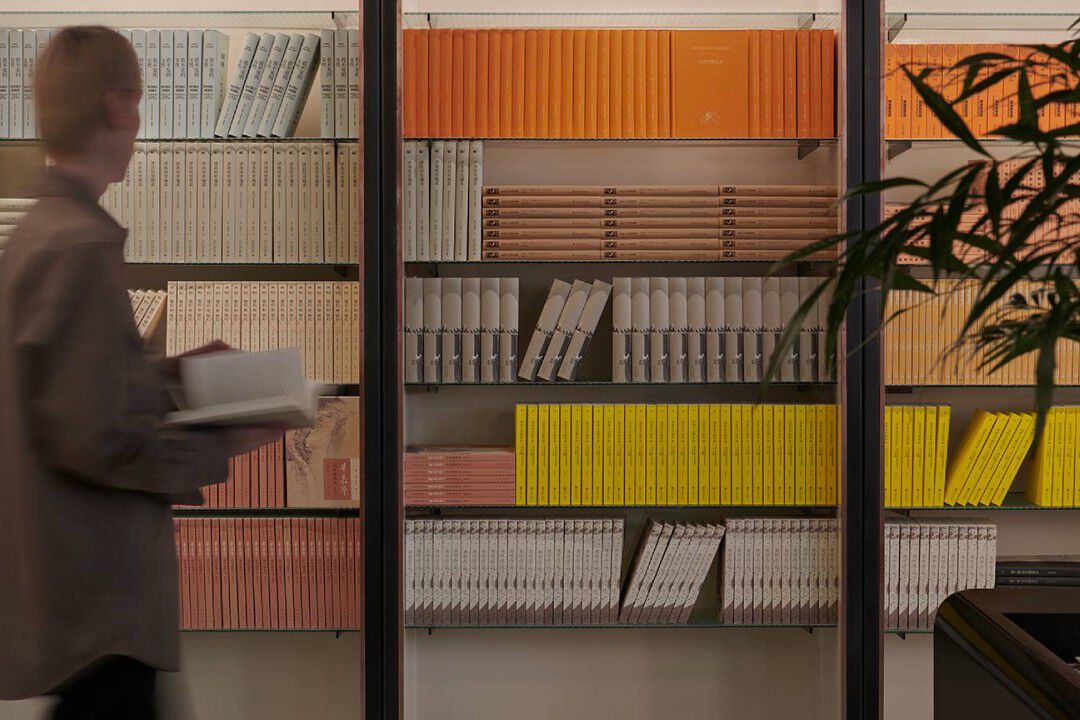 Blurred person walking past warm wooden shelves filled with yellow, orange, and red books.