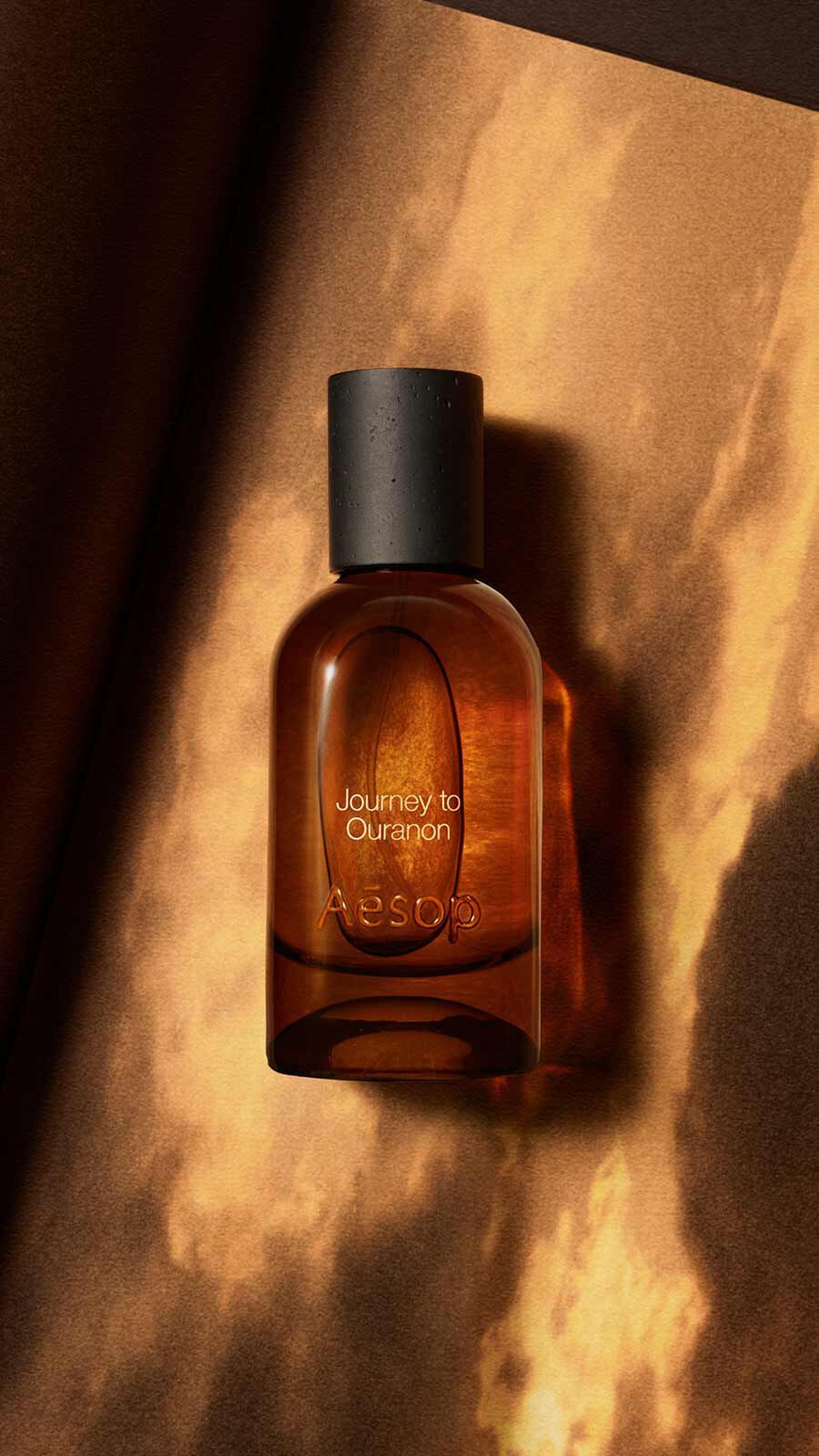 Aesop Journey to Ouranon Eau de Parfum bottle with black cap against a dramatic warm orange and golden cloud sky.