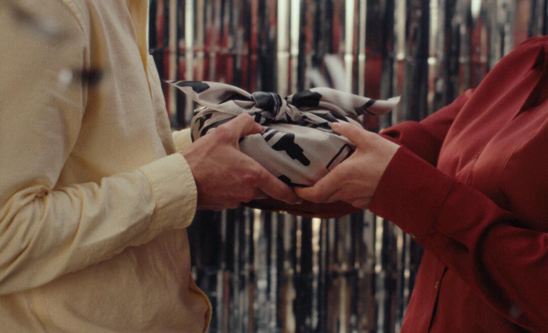 Two people holding a gift wrapped in patterned fabric between them, wearing cream and red shirts, against a silver tinsel backdrop.
