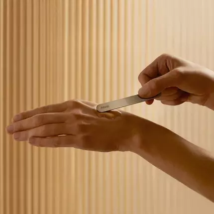 A hand uses an Aesop spatula to apply skincare serum to another hand.