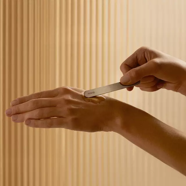 A hand uses an Aesop spatula to apply skincare serum to another hand.