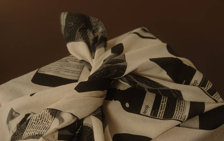A close-up photograph of a package wrapped in a cream-colored cotton fabric using a traditional knotting technique (Furoshiki). The fabric is patterned with black-and-white graphics of Aesop bottles and text labels. The package is set against a warm, minimalist brown background under soft lighting.