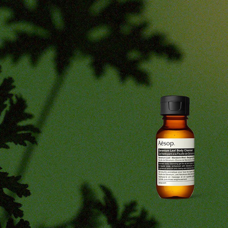 An amber glass bottle of Aesop Geranium Leaf Body Cleanser on a textured green background, with soft shadows of plant leaves on the left.