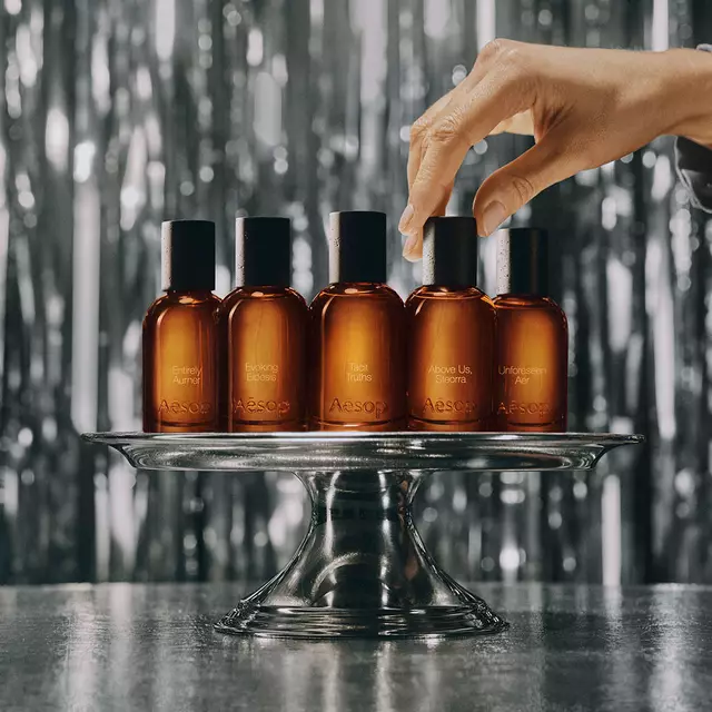 Five Aesop fragrance bottles on a glass stand as a hand reaches for one.
