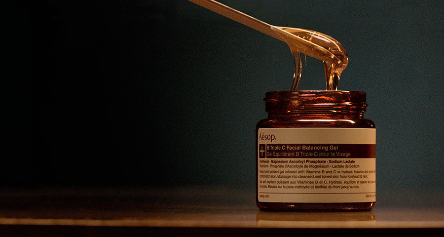 A golden, viscous gel is being scooped from an amber Aesop jar with a golden spoon, set against a dark background.