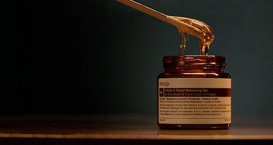 A golden, viscous gel is being scooped from an amber Aesop jar with a golden spoon, set against a dark background.
