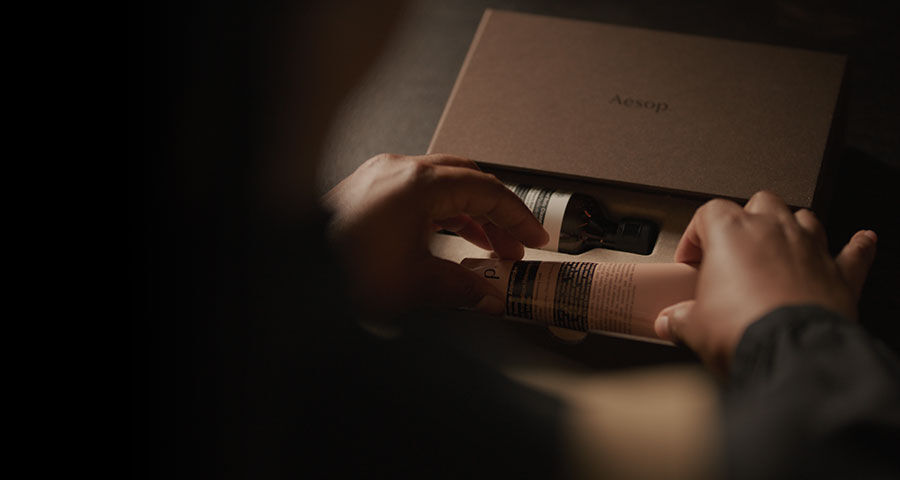 A person's hands are carefully removing a beige-colored tube and a dark bottle, both cosmetic products, from a brown cardboard box.