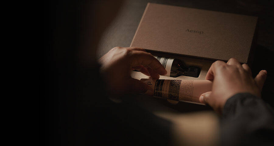 A person's hands are carefully removing a beige-colored tube and a dark bottle, both cosmetic products, from a brown cardboard box.