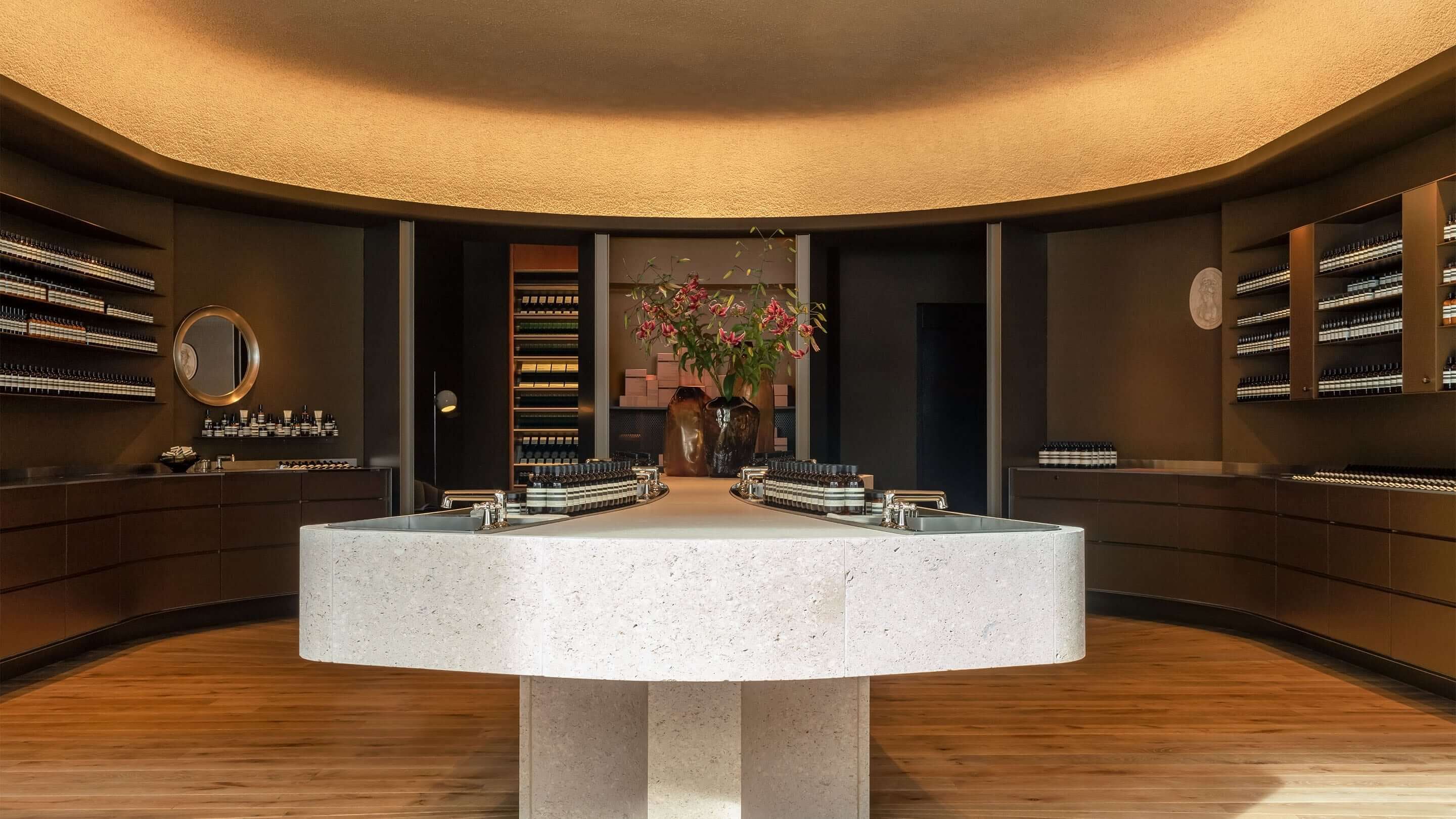 An elegant beauty boutique with minimalist design, featuring a large stone central island with integrated sinks and products, surrounded by dark wood shelving and warm ambient lighting.