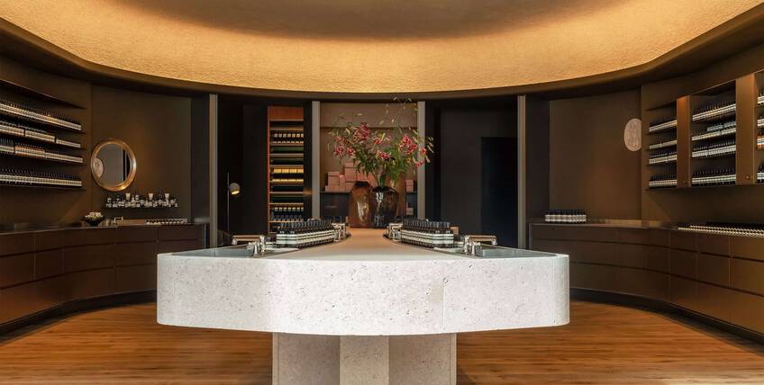 An elegant beauty boutique with minimalist design, featuring a large stone central island with integrated sinks and products, surrounded by dark wood shelving and warm ambient lighting.