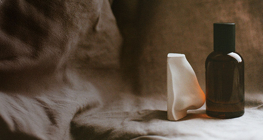 A dark amber bottle with a black cap sits next to a white, sculptural nose-like object on a rumpled fabric surface.