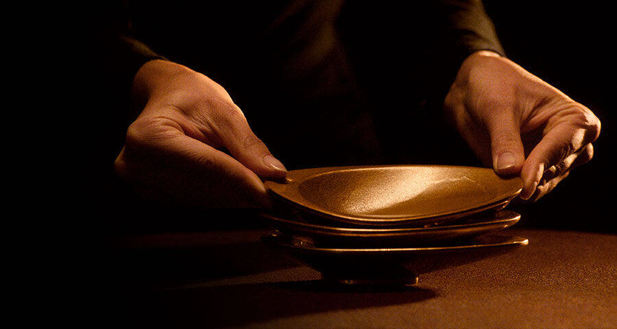 Two hands are carefully placing a golden, shallow dish on top of a stack of similar dishes, against a dark background with warm, subtle lighting.
