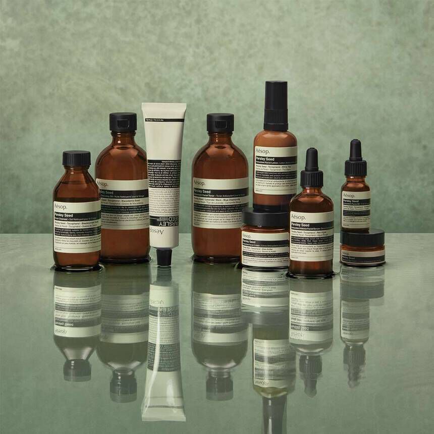 Amber Aesop skincare bottles, jars, and dropper serums arranged on a reflective surface against a muted green background.